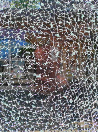 A close-up view of shattered glass reveals a distorted reflection of a nearby storefront, with sunlight casting intricate patterns through the cracks