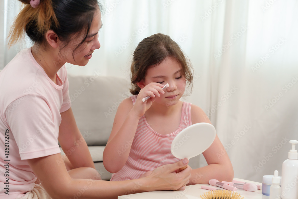 Caucasian girl learning makeup with Asian mom. Little girl wearing pink sleeveless top using brush while looking into handheld mirror. Mother sitting beside, watching with gentle smile in home setting Caucasian girl learning makeup with Asian mom. Little girl wearing pink sleeveless top using brush while looking into handheld mirror. Mother sitting beside, watching with gentle smile in home setting