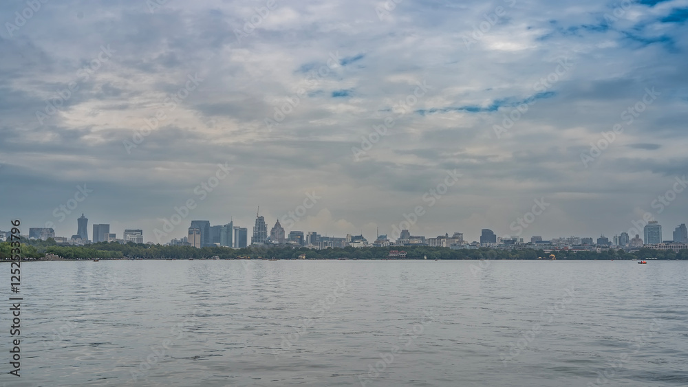 Beautiful calm lake. Ripples on the surface of the water. In the distance, there is green vegetation on the shore. Tall city buildings, skyscrapers against a blue sky and clouds. China. Hangzhou. Beautiful calm lake. Ripples on the surface of the water. In the distance, there is green vegetation on the shore. Tall city buildings, skyscrapers against a blue sky and clouds. China. Hangzhou.