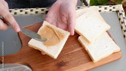 Male hands spreading peanuts butter on loaf of bread above table.