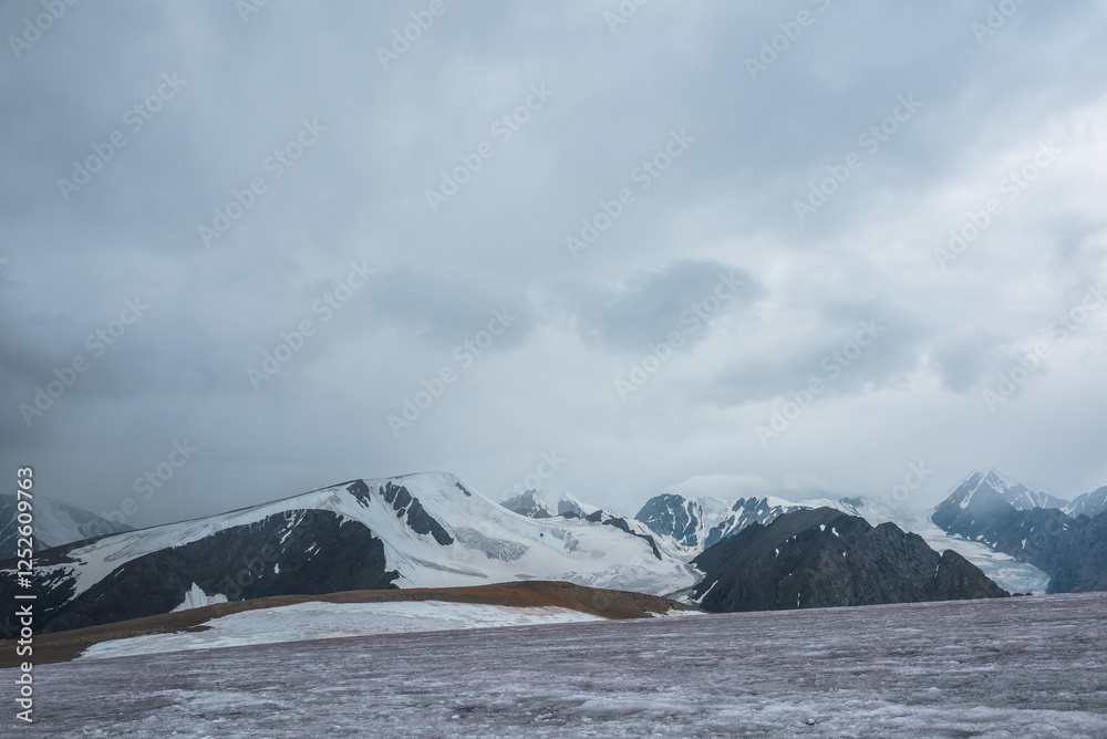 Dramatic vast misty top view from ice pass to big glacier tongue among sharp rocks and large snow-capped mountains in rainy low clouds. Dark atmospheric mountain silhouettes in rain in gray cloudy sky Dramatic vast misty top view from ice pass to big glacier tongue among sharp rocks and large snow-capped mountains in rainy low clouds. Dark atmospheric mountain silhouettes in rain in gray cloudy sky