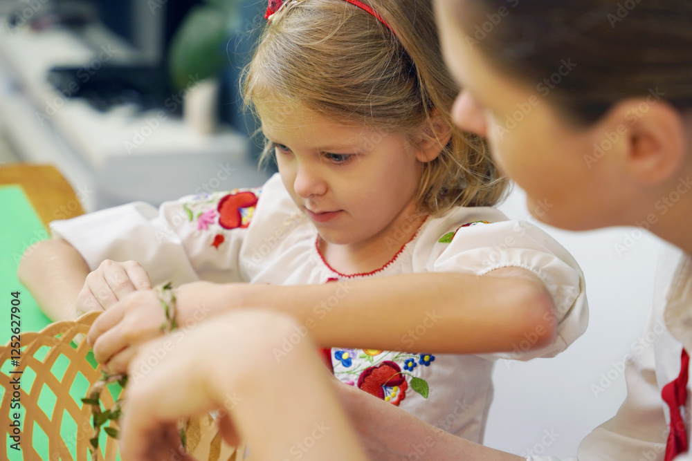 Obraz premium Mother and daughter at the table in traditional Ukrainian costumes weaving a basket for Easter
