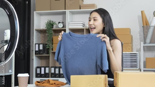 An Asian female business owner records a live vlog, showing a parcel box in front of a smartphone. She sells clothes online, engaging customers and promoting products for successful e-commerce sales