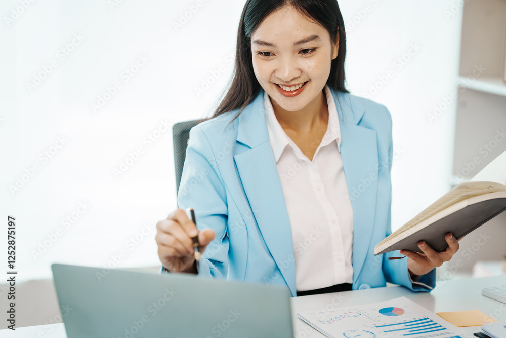 An Asian female accountant works happily at her desk in the office, using a laptop to analyze financial charts. She manages business finance and online transactions for company success An Asian female accountant works happily at her desk in the office, using a laptop to analyze financial charts. She manages business finance and online transactions for company success
