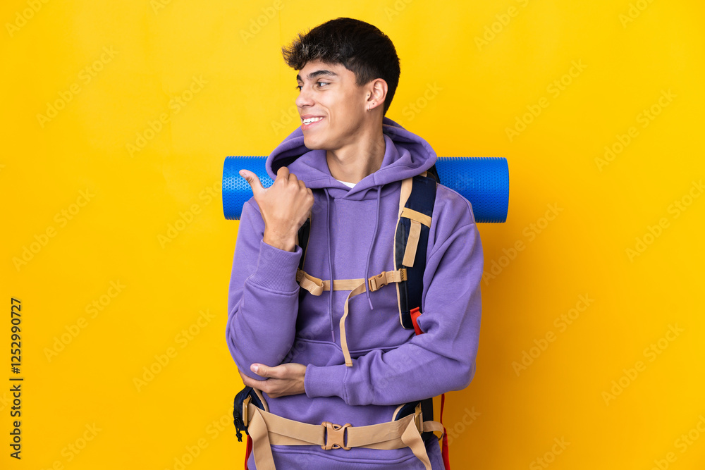 Young mountaineer man with a big backpack over isolated yellow background pointing to the side to present a product Young mountaineer man with a big backpack over isolated yellow background pointing to the side to present a product