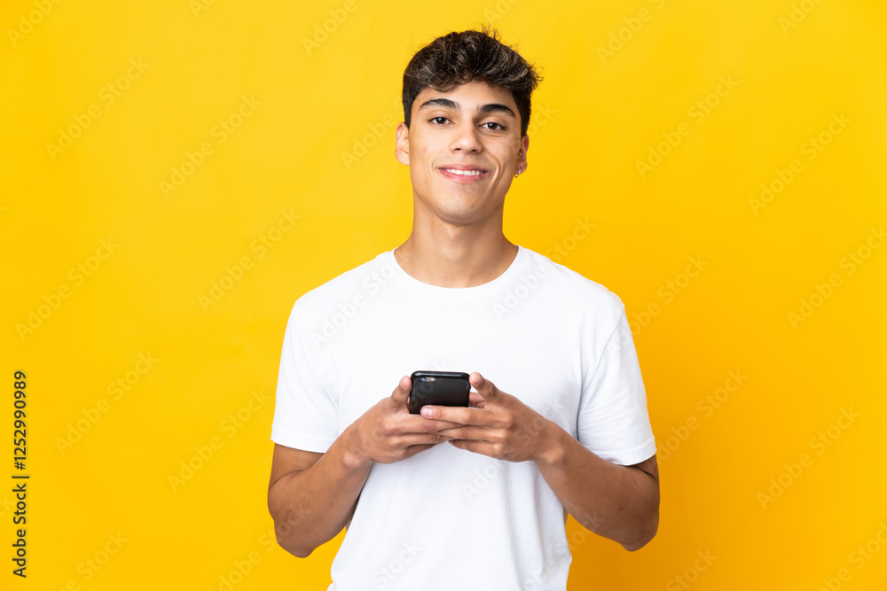 Young man over isolated yellow background looking at the camera and smiling while using the mobile Young man over isolated yellow background looking at the camera and smiling while using the mobile