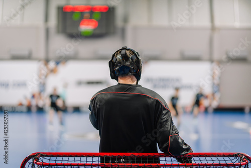 Принти на полотні A floorball goalkeeper wearing a black jersey and helmet stands behind the net, focused on the game, with a blurred scoreboard in the background