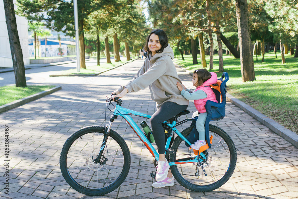 Mother riding bicycle with her young daughter sitting in a child seat on the back. They are enjoying a sunny day in green park with paved pathways, surrounded by trees and nature. Mother riding bicycle with her young daughter sitting in a child seat on the back. They are enjoying a sunny day in green park with paved pathways, surrounded by trees and nature.