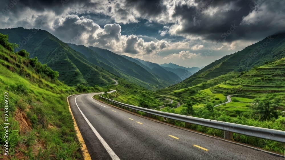 Naklejka premium A winding mountain road through lush green valleys under a dramatic cloudy sky.