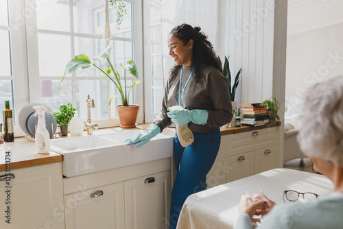 Rare view of elderly lonely woman talking to young african american female helping volunteer while she cleaning her kitchen sink in blue raisin gloves, spraying and mopping dirt from surfaces