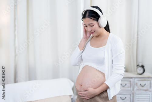 Beautiful pregnant Asian woman nearing birth. Smiling while wearing headphones and gently holding belly. Dressed in a white top and cardigan with beige pants. Cozy home interior with soft lighting.