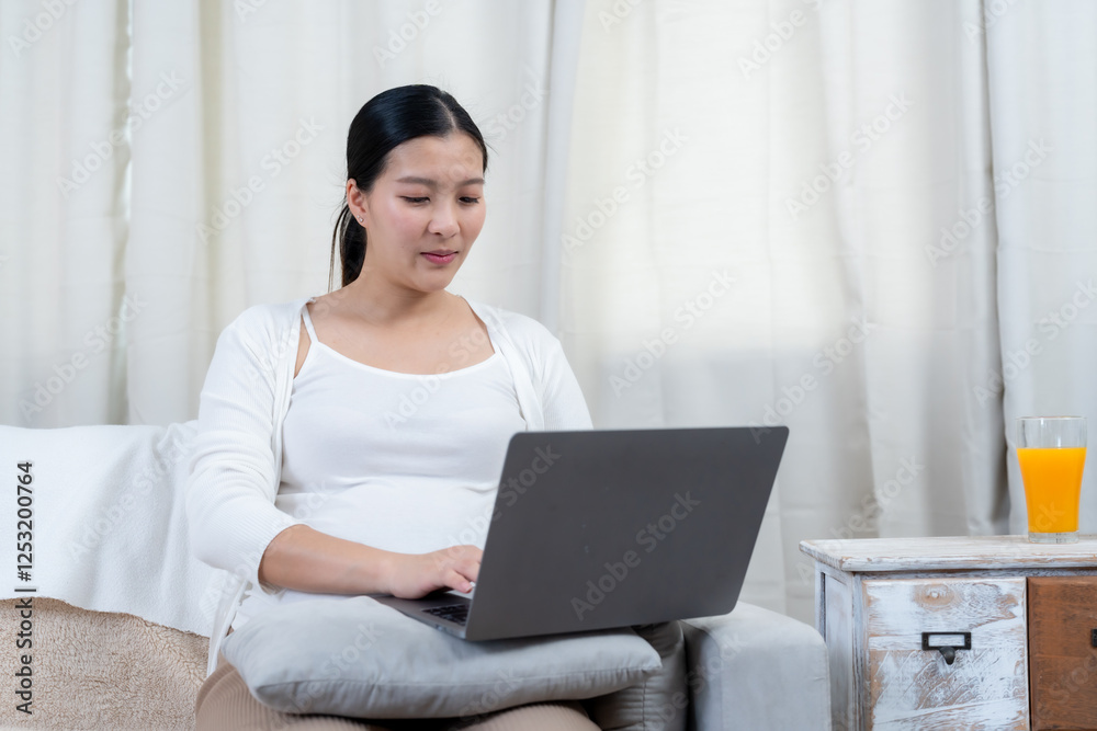 Beautiful pregnant Asian woman nearing birth. Using a laptop while sitting comfortably on a soft couch. Wearing a white top and cardigan with beige pants. Cozy home setting with natural light Beautiful pregnant Asian woman nearing birth. Using a laptop while sitting comfortably on a soft couch. Wearing a white top and cardigan with beige pants. Cozy home setting with natural light