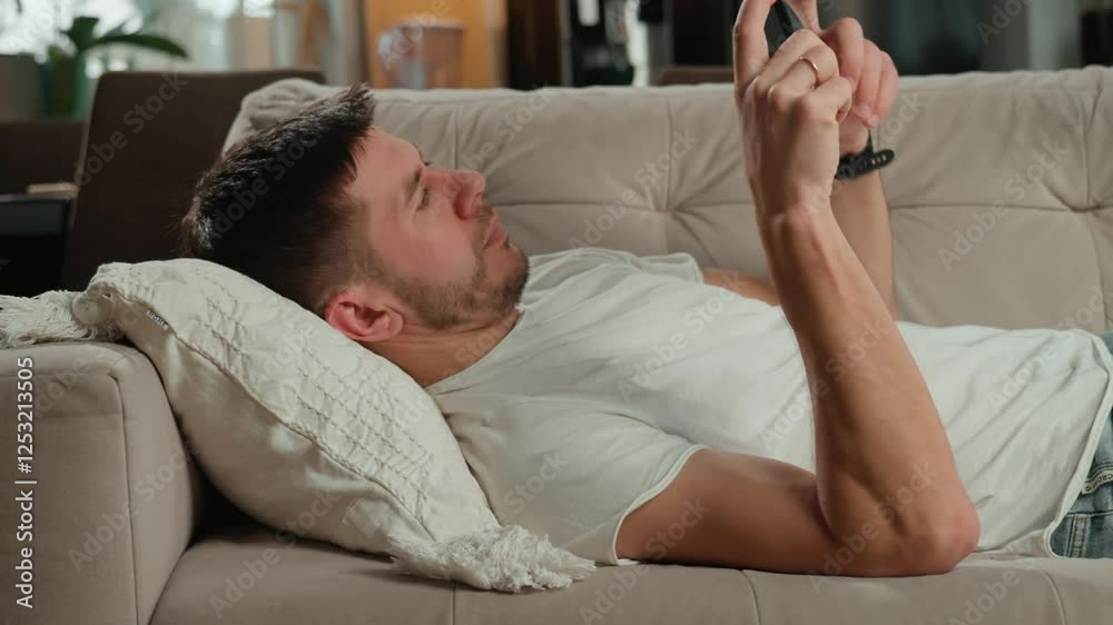Man in white t-shirt and jeans relaxing on beige sofa, using his smartphone in living room. Male resting on couch browsing social media on mobile phone Man in white t-shirt and jeans relaxing on beige sofa, using his smartphone in living room. Male resting on couch browsing social media on mobile phone