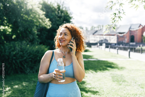 Portrait of curly-haired ginger head woman calling to her husband to take her from yoga training outdoor standing on lawn in fitness clothes, carrying sport bag and holding bottle of water