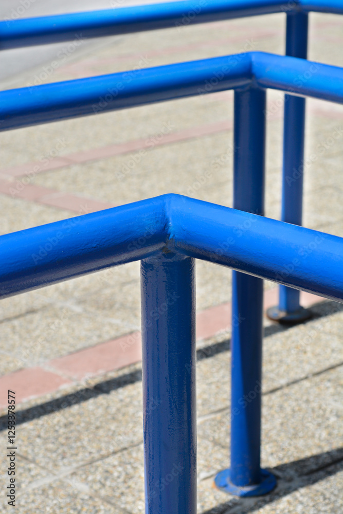 A close-up shot of a bright blue metal railing with a blurred background of a paved surface. A close-up shot of a bright blue metal railing with a blurred background of a paved surface.