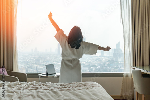 Hotel relaxation on lazy day with Asian woman waking up from good sleep on bed in weekend morning resting in comfort bedroom looking toward city view, having happy work-life quality balance lifestyle