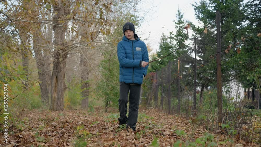 Young boy in blue jacket walking along autumn path holding stick in hand scattering the dry leaves on the ground, surrounded by dry leaves, playfully interacting with nature in forest Young boy in blue jacket walking along autumn path holding stick in hand scattering the dry leaves on the ground, surrounded by dry leaves, playfully interacting with nature in forest