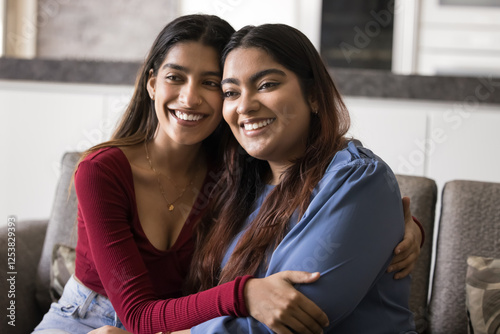 Two attractive Indian women sitting close together on sofa in cozy modern living room, cuddling, feeling sibling love, showing mutual support, enjoy shared moments. Closeness and trust between sisters