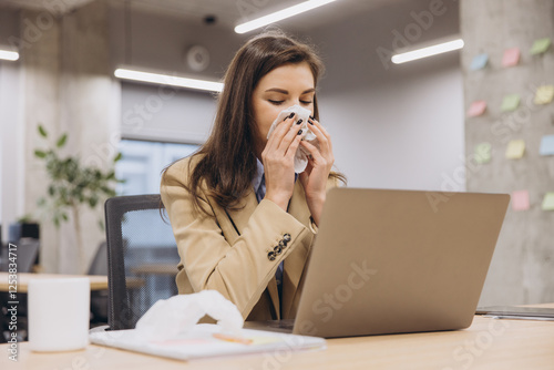 Professional businesswoman experiencing cold symptoms, wiping nose while working on laptop at clean, organized desk during office hours