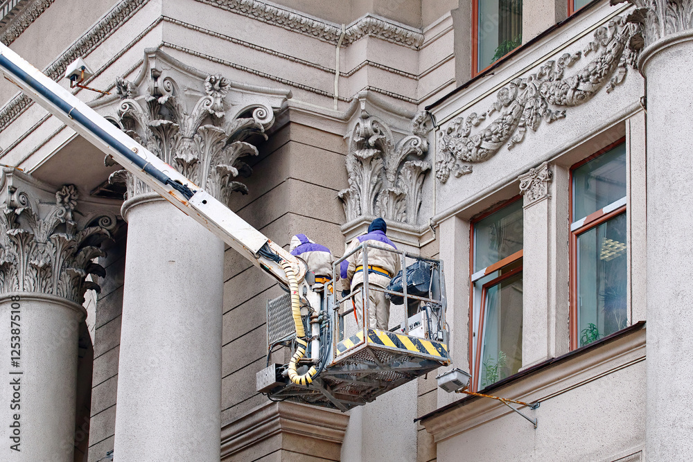 Workers on lift platform paint facade, facade restoration on historic building with ornate columns, preservation of urban architecture. Men on lift platform clean and maintain historic building