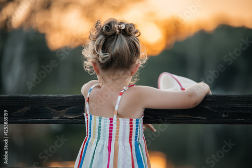 A small girl about 5 years old standing with her back to the camera, looking at a warm sunset horizon. Light summer dress, golden hour lighting, calm atmospheric mood, concept of childhood, dreaminess