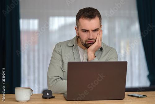 Bored man working overtime, sitting at the desk in front of a laptop and annoyed looking on the screen.