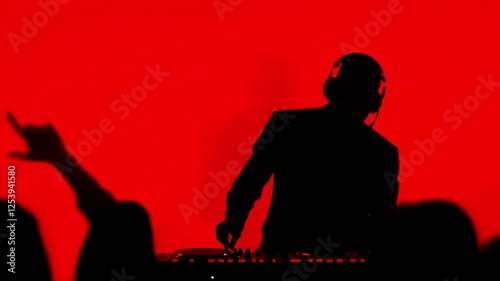 Silluette of a DJ playing music, red monotone background as the DJ switches knobs on the mixer and the crowd dancing