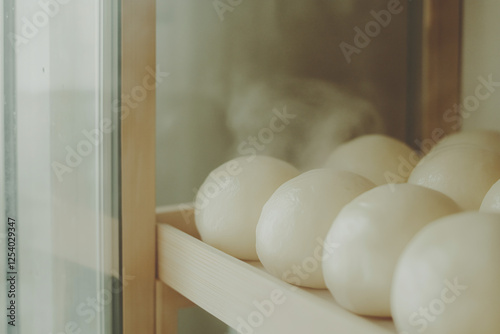 Steamed Buns Rising: Soft, pillowy steamed buns rest on a wooden shelf within a glass-fronted warming cabinet, emitting a gentle steam, a picture of comforting warmth and delicious anticipation. photo