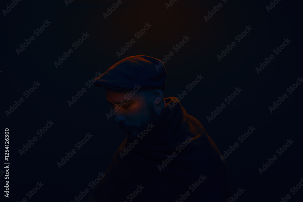 Portrait of a man with his head bowed, wrapped in a dark scarf, wearing a cap with a subtle orange glow illuminating parts of the face. Portrait of a man with his head bowed, wrapped in a dark scarf, wearing a cap with a subtle orange glow illuminating parts of the face.