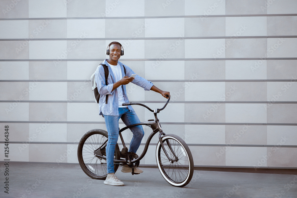 Black male hipster with mobile gadget and headphones riding bicycle near brick wall on urban street, free space Black male hipster with mobile gadget and headphones riding bicycle near brick wall on urban street, free space