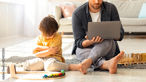 Family issues because of work. Upset little boy sitting offended with toys on floor, busy concentrated father working on laptop from home, sitting near son, free space photo