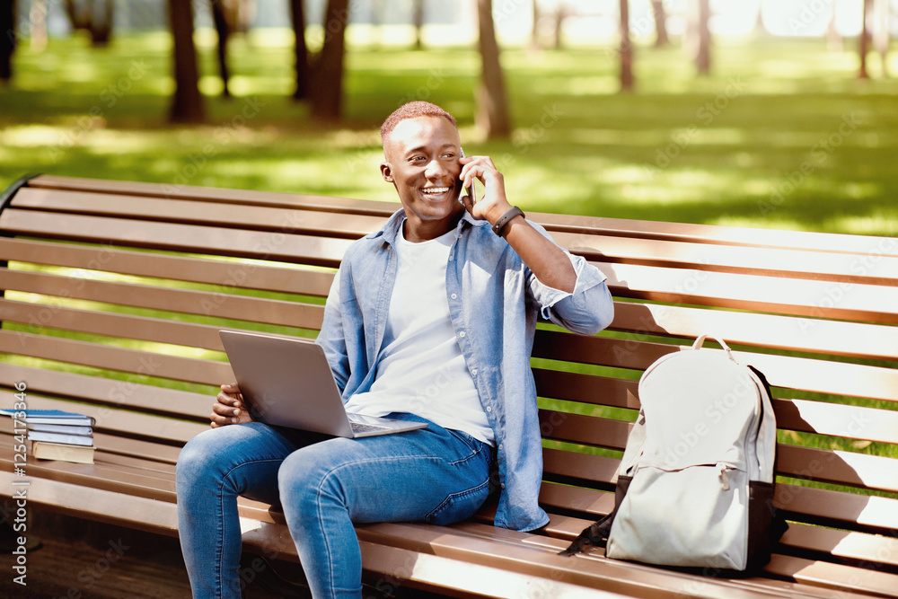 Fototapeta premium E-learning concept. Funky black student with laptop talking on smartphone on bench at park. African American guy talking to his college tutor while watching educational video on pc, outside