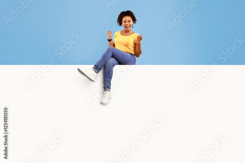 Beautiful millennial African American lady making YES gesture, sitting on top of blank paper banner, excited over huge sale or promo, offering mockup for your advertisement on blue studio background photo