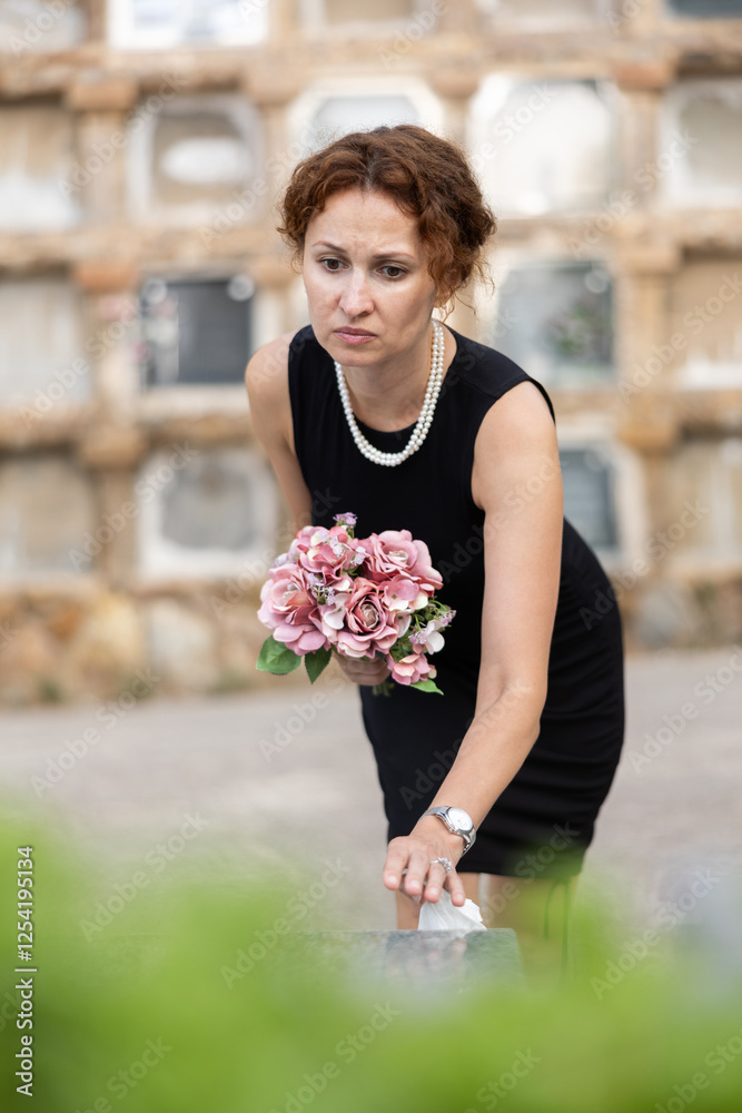Fototapeta premium Sad moment of visiting cemetery and worshiping memory of deceased relative. Thoughtful and sorrowful woman puts bouquet of hydrangeas on grave of deceased friend, mourns loss