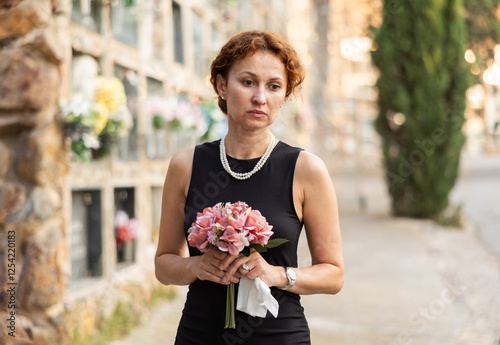 Sorrowful young woman in black dress with bouquet of flowers and handkerchief in hands, stands in cemetery. Visiting grave of deceased relative, mourning dead loved one. Appeal to God, prayer for dead photo