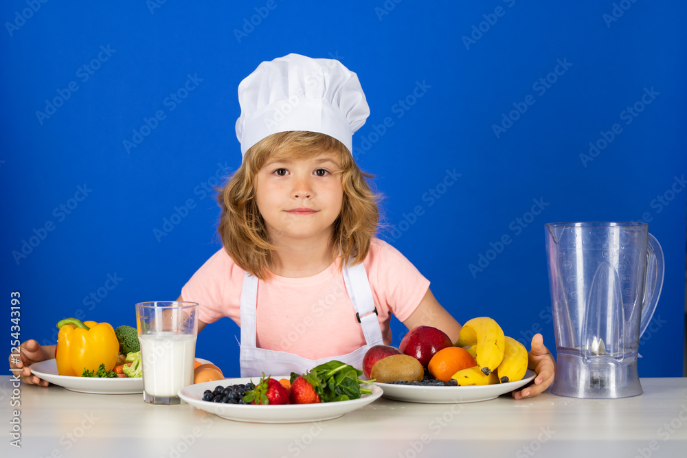 Child chef cook prepares food in isolated blue studio background. Kids cooking. Teen boy with apron and chef hat preparing a healthy vegetables meal in the kitchen. Child chef cook prepares food in isolated blue studio background. Kids cooking. Teen boy with apron and chef hat preparing a healthy vegetables meal in the kitchen.