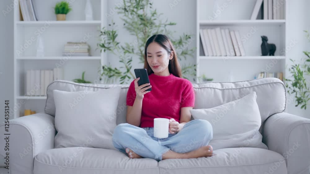 Attractive smiling young Asian woman using mobile phone while sitting on the sofa at home Attractive smiling young Asian woman using mobile phone while sitting on the sofa at home