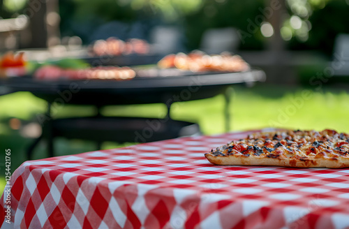 Delicious pizza on a red and white checkered tablecloth, with a barbecue grill featuring various meats grilling outdoors during summer season photo