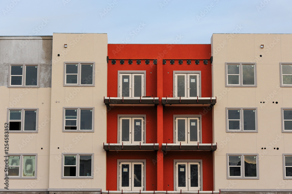 Fototapeta premium A modern apartment building under construction featuring a mix of beige and red exterior walls, multiple balconies, and large windows as part of an urban residential development proj