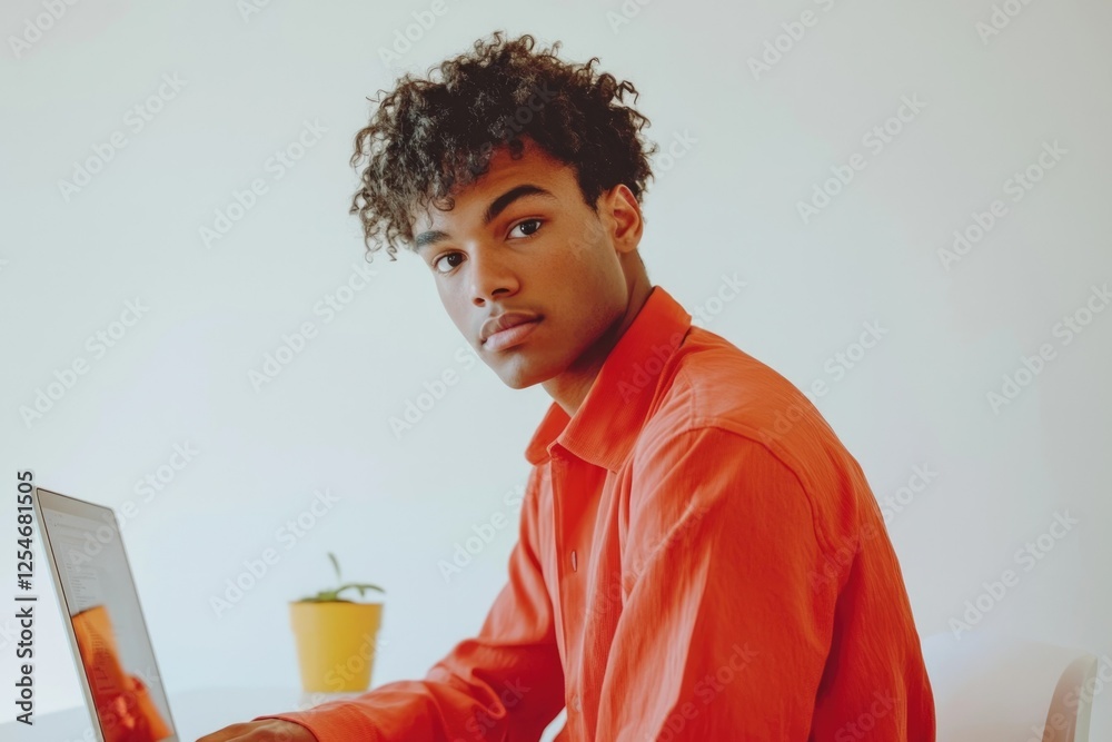 Young man with curly hair, wearing an orange shirt, works on a laptop. Young man with curly hair, wearing an orange shirt, works on a laptop.