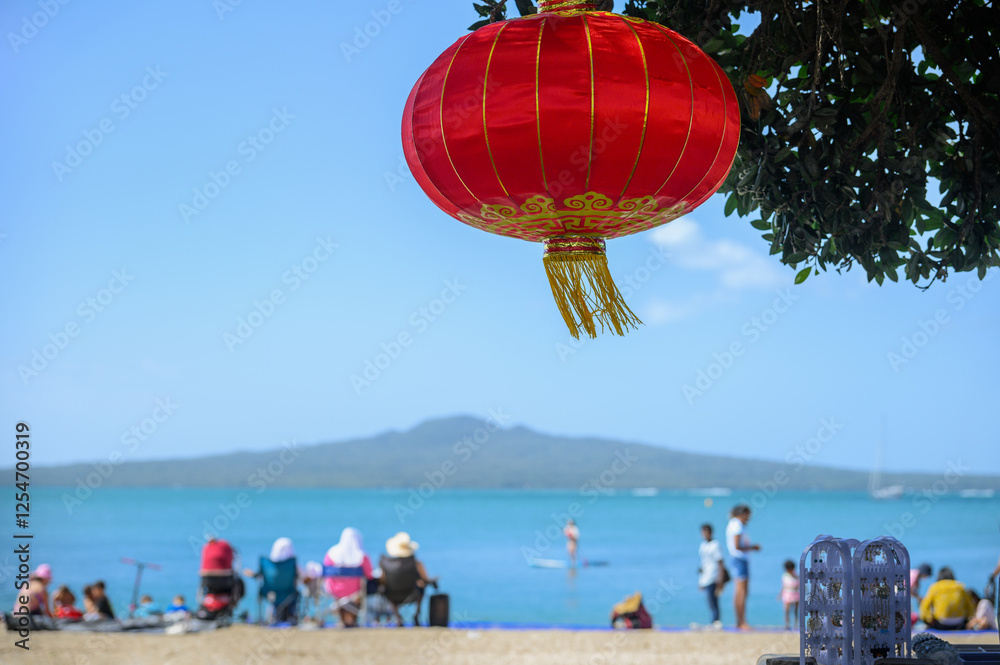 Red lanterns hanging under the Pohutukawa trees. Unrecognizable crowd of people enjoying summer at Mission Bay. Auckland.