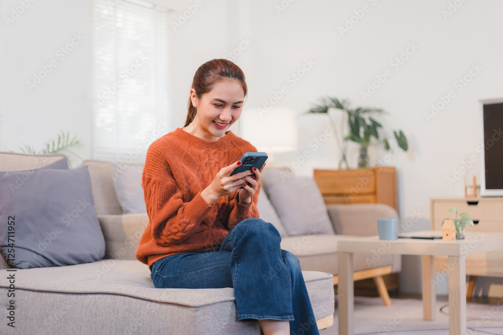 Woman in cozy orange sweater sits on sofa, smiling while using her smartphone in bright living room. Woman in cozy orange sweater sits on sofa, smiling while using her smartphone in bright living room.