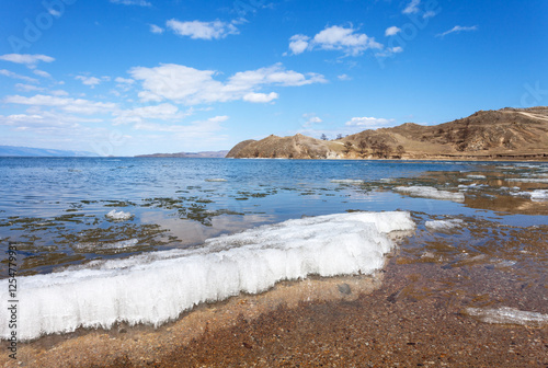 Beautiful bright spring landscape with melting ice on the sandy beach of Baikal Lake on a sunny day. View of Small Sea Strait from the Kurkut Bay. Outdoor recreation on lake shore. Natural background photo