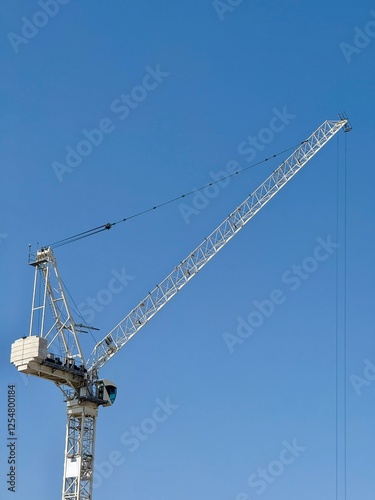 A tall tower crane with a long boom extending against a clear blue sky, used for heavy lifting at construction sites. The operator's cabin and counterweights are visible.