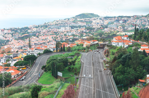 Scenic view of Madeira, highway with multiple lanes and several cars driving, surrounded by lush greenery and residential buildings with red tiled roofs. In background, there is a Funchal city