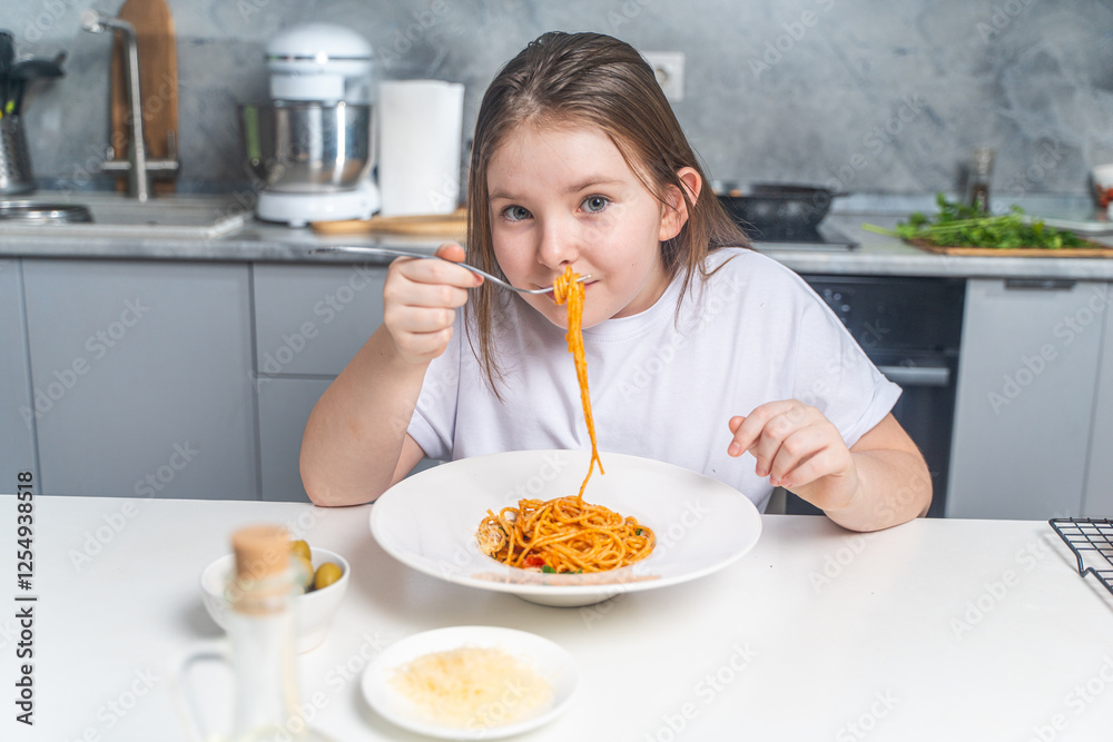 Cute little teen girl eating spaghetti on the background of gray kitchen at home sitting in white t-shirt at table.