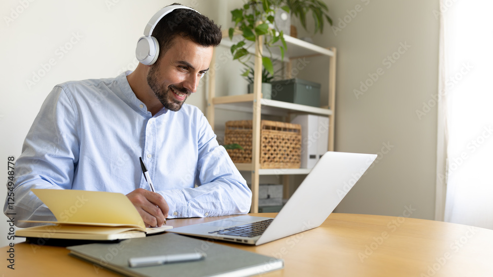 Obraz premium Happy man with headphones writing notes working at home using laptop. Copy space.