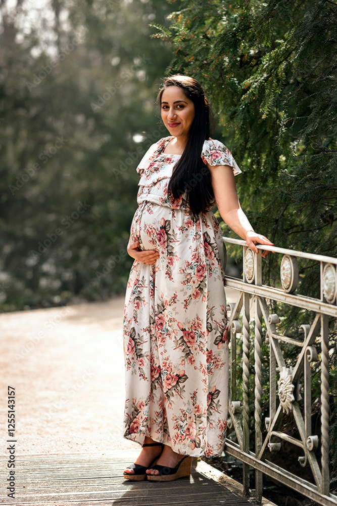 Portrait of pregnant woman with long black hair in park dressed in white dress with flowers, woman holds her baby belly, pregnant woman in stands in park, indian woman portrait Portrait of pregnant woman with long black hair in park dressed in white dress with flowers, woman holds her baby belly, pregnant woman in stands in park, indian woman portrait
