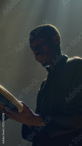 Vertical low angle view of Black pastor wearing dark cassock carefully studying bible book in catholic church photo