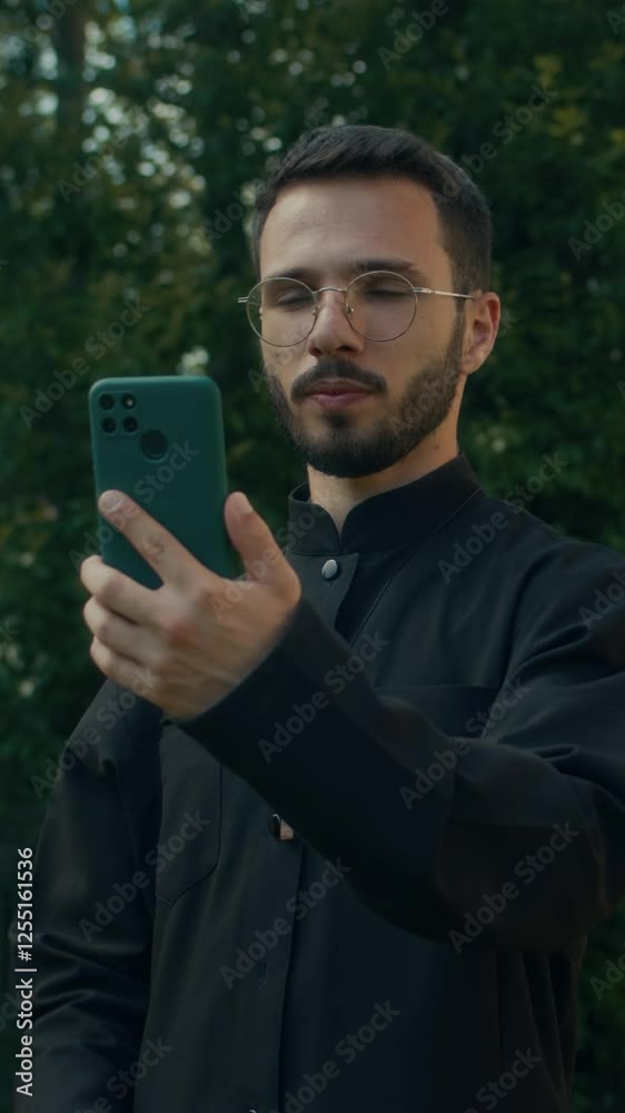 Vertical view of bearded Middle Eastern young pastor conducting online consultation using smartphone with green trees in background Vertical view of bearded Middle Eastern young pastor conducting online consultation using smartphone with green trees in background